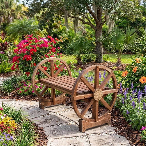 A rustic wooden bench with decorative wheels on the sides, situated on a stone pathway in a lush, colorful garden with flowers and trees.