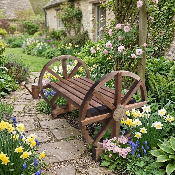 A rustic garden bench built with two decorative wooden wheels on the sides, set in a lush, flower-filled stone path garden.