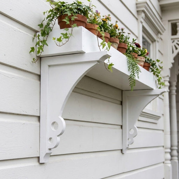 White decorative wall-mounted shelf brackets holding terracotta pots with green ivy and flowers, attached to a white house.