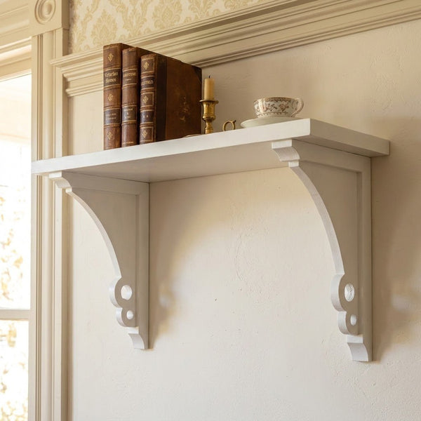 A white decorative wall shelf mounted on a cream-colored wall, with three brown books, a brass candlestick, and a teacup.