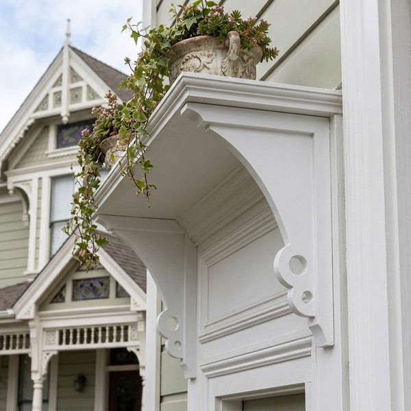 White decorative architectural brackets corbels on the exterior of a house, with potted green plants resting on top.