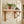 A light natural wood wall-mounted shelf with decorative bracket supports, styled with several potted green plants against a white brick wall next to a window.