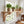 A light natural wood wall-mounted shelf with decorative corset brackets is installed above a white window in a bright kitchen. Several potted herbs and small plants sit on the shelf, along with a small brass oil lamp and a wooden cutting board with a mortar and pestle below.