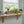 A rustic wooden floating shelf mounted on a tiled wall under a window, styled with a rectangular planter box containing green plants, two rolled gray towels, and three amber glass bottles.