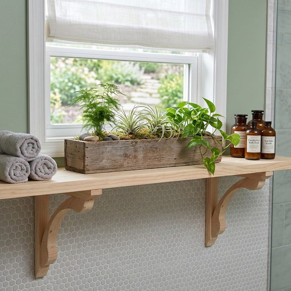 A rustic wooden floating shelf mounted on a tiled wall under a window, styled with a rectangular planter box containing green plants, two rolled gray towels, and three amber glass bottles.