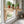 Two light natural wood wall-mounted shelf brackets installed on a white kitchen window, holding several potted green herbs and small plants.