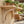 A light natural wood wall-mounted shelf with decorative corset brackets, shown attached to a beige stucco wall in an outdoor patio setting with potted plants and wooden chairs visible in the background.