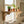 A light natural wood wall-mounted shelf with decorative corset brackets is installed beneath a window in a bright bathroom. Several small potted houseplants sit on the shelf, and a folded towel rests on the windowsill.