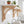 Three light natural wood corset wall brackets holding a white shelf in a bright, white-tiled bathroom. The shelf is styled with potted green plants, and a chrome faucet and folded towels are visible below.