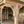 A close-up of a wooden porch support post and sunburst bracket under a roof overhang, with a front door, deck flooring, a potted fern, and a rocking chair visible in the background.